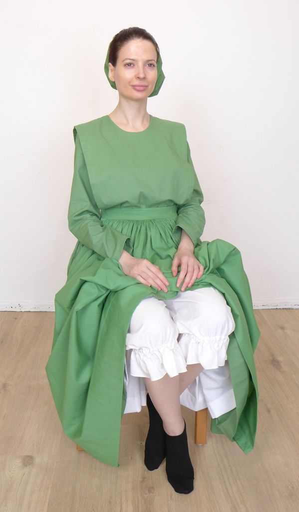 Front view of a woman in a plain cape dress, sitting on a chair, showing her undergarments